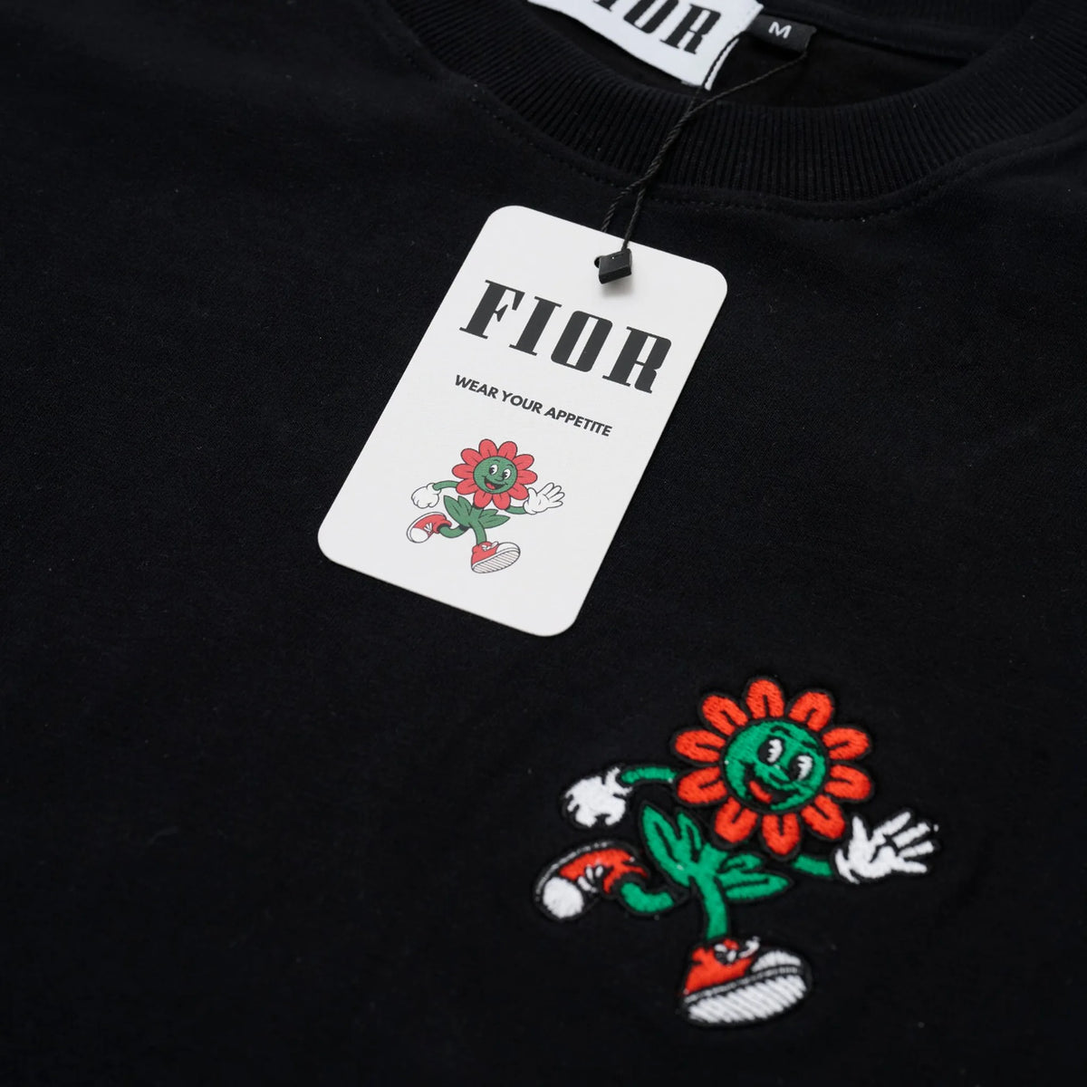 Cartellino FIOR e ricamo mascotte su T-shirt nera, dettaglio materiale e brand.